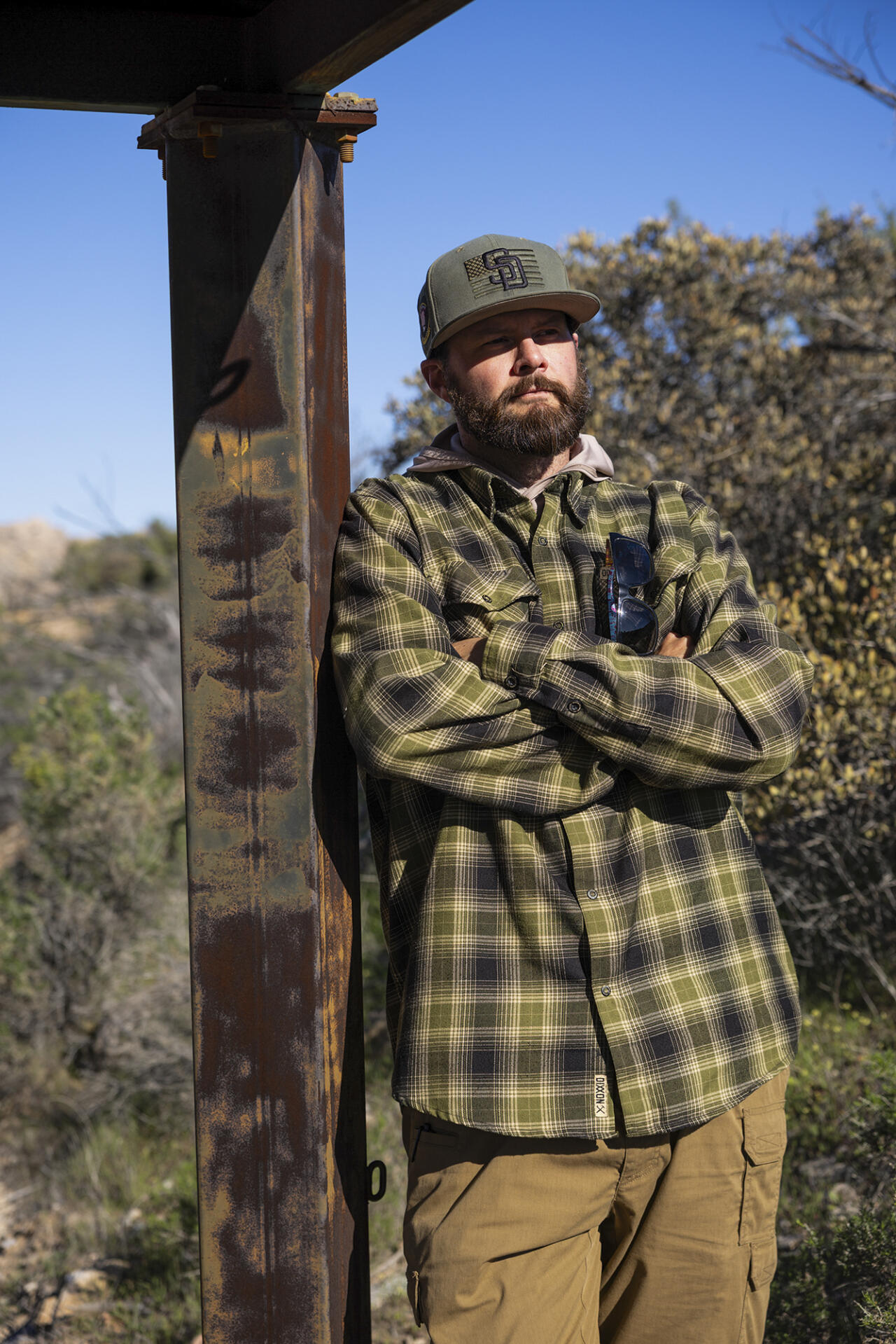A person wearing a baseball cap and plaid shirt stands with arms crossed beside a rusted metal post outdoors, with shrubs and a clear blue sky in the background.