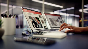 A close view shows a person using a laptop displaying a news website with a headline and photo on the screen. Another laptop sits nearby on the desk, and the scene is set in a modern office environment with glass walls and overhead lighting.