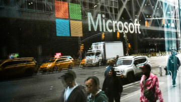 The reflection of people walking along a sidewalk is seen on a glass window of a storefront with the Microsoft logo on it. Yellow taxis, trucks and cars can also be seen in the reflection.