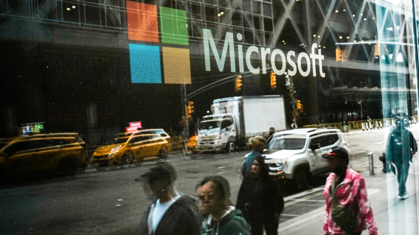 The reflection of people walking along a sidewalk is seen on a glass window of a storefront with the Microsoft logo on it. Yellow taxis, trucks and cars can also be seen in the reflection.