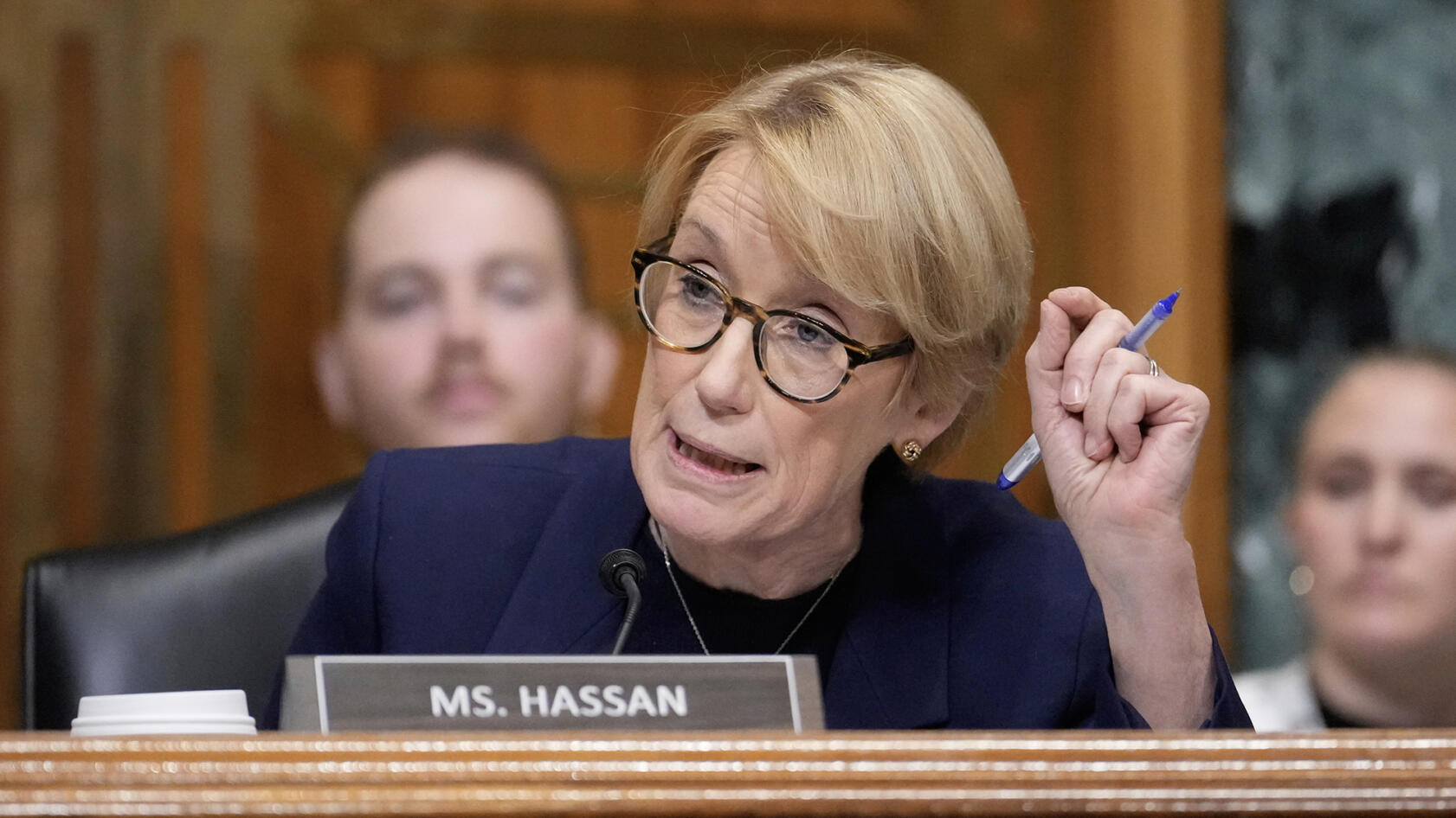 A woman wearing glasses speaks into a microphone during a congressional hearing while holding a blue pen in one hand. A nameplate reading “MS. HASSAN” sits in front of her as other attendees appear out of focus in the background.