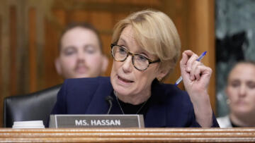 A woman wearing glasses speaks into a microphone during a congressional hearing while holding a blue pen in one hand. A nameplate reading “MS. HASSAN” sits in front of her as other attendees appear out of focus in the background.