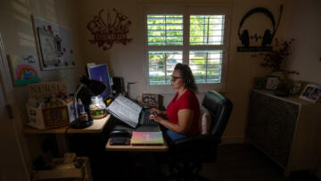 A person wearing a red shirt and a headset sits in front of a desk and types into a keyboard, in a dimly lit room with a window overlooking a residential street.