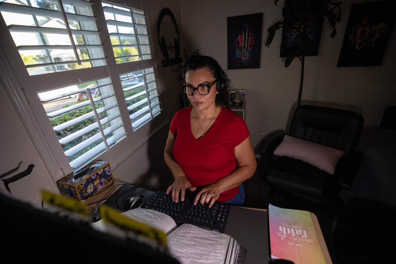 A person wearing a red shirt and a headset sits in front of a desk and types into a keyboard, in a dimly lit room with a window overlooking a residential street.