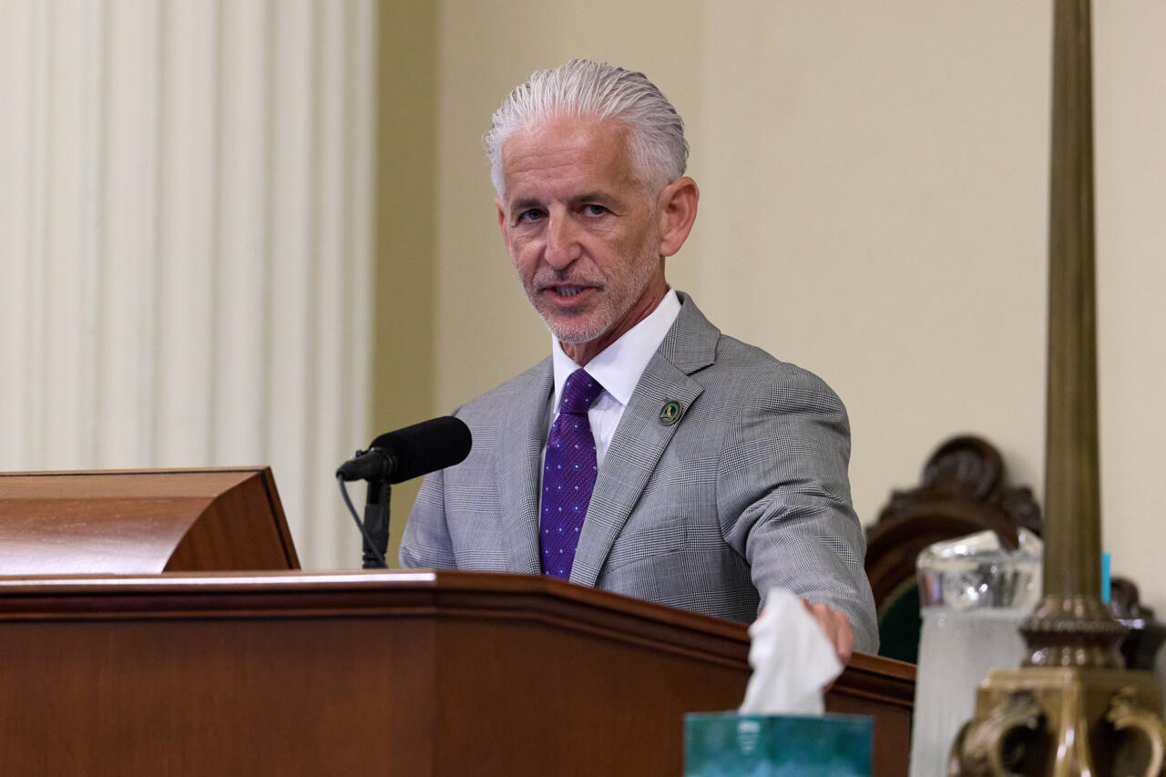 A person, with white hair and wearing a gray suit, stands in front of a podium with a microphone on it while they speak.