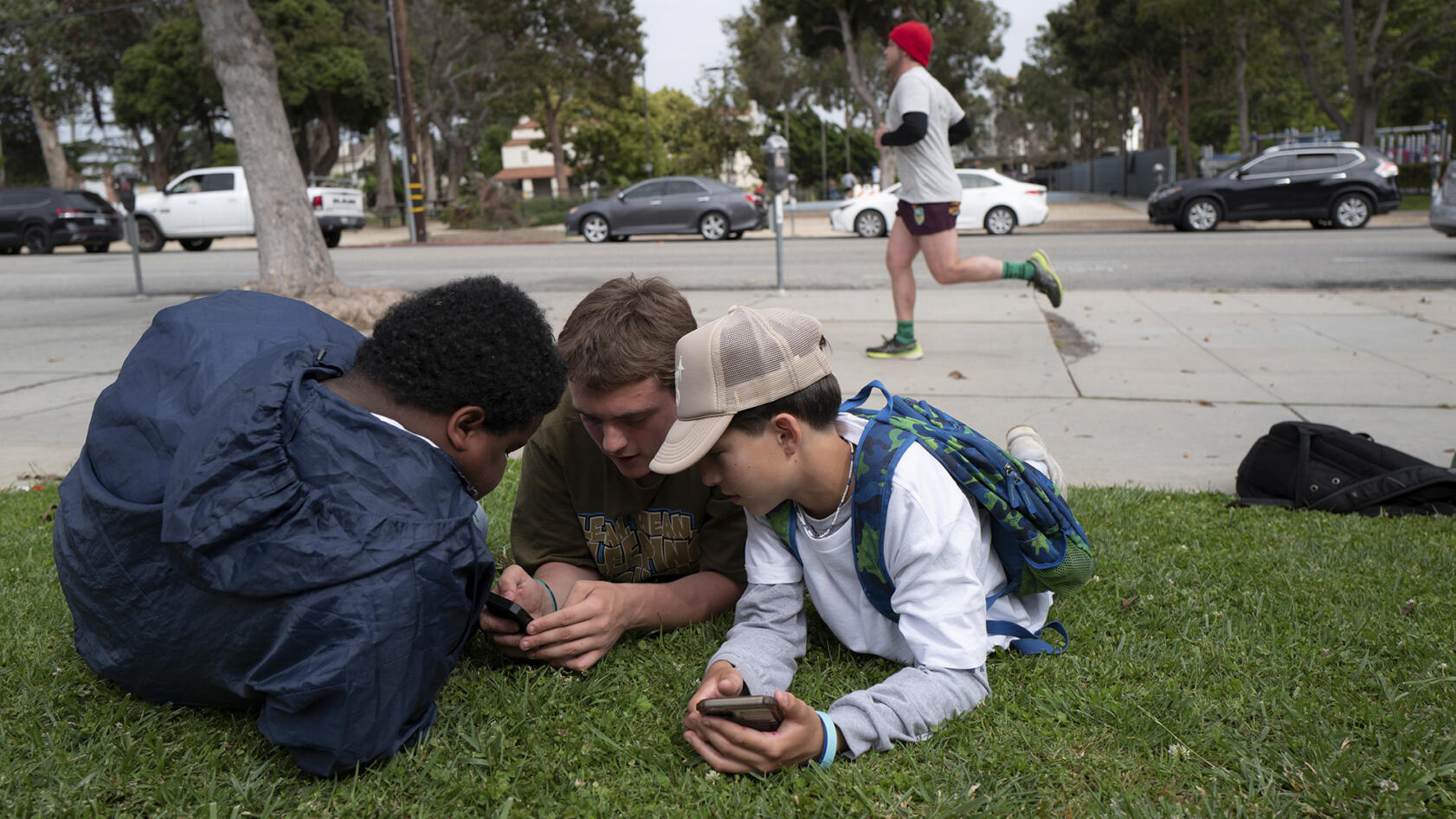 A group of students lying in the grass near a sidewalk, looking at a cellphone while a man runs past them.