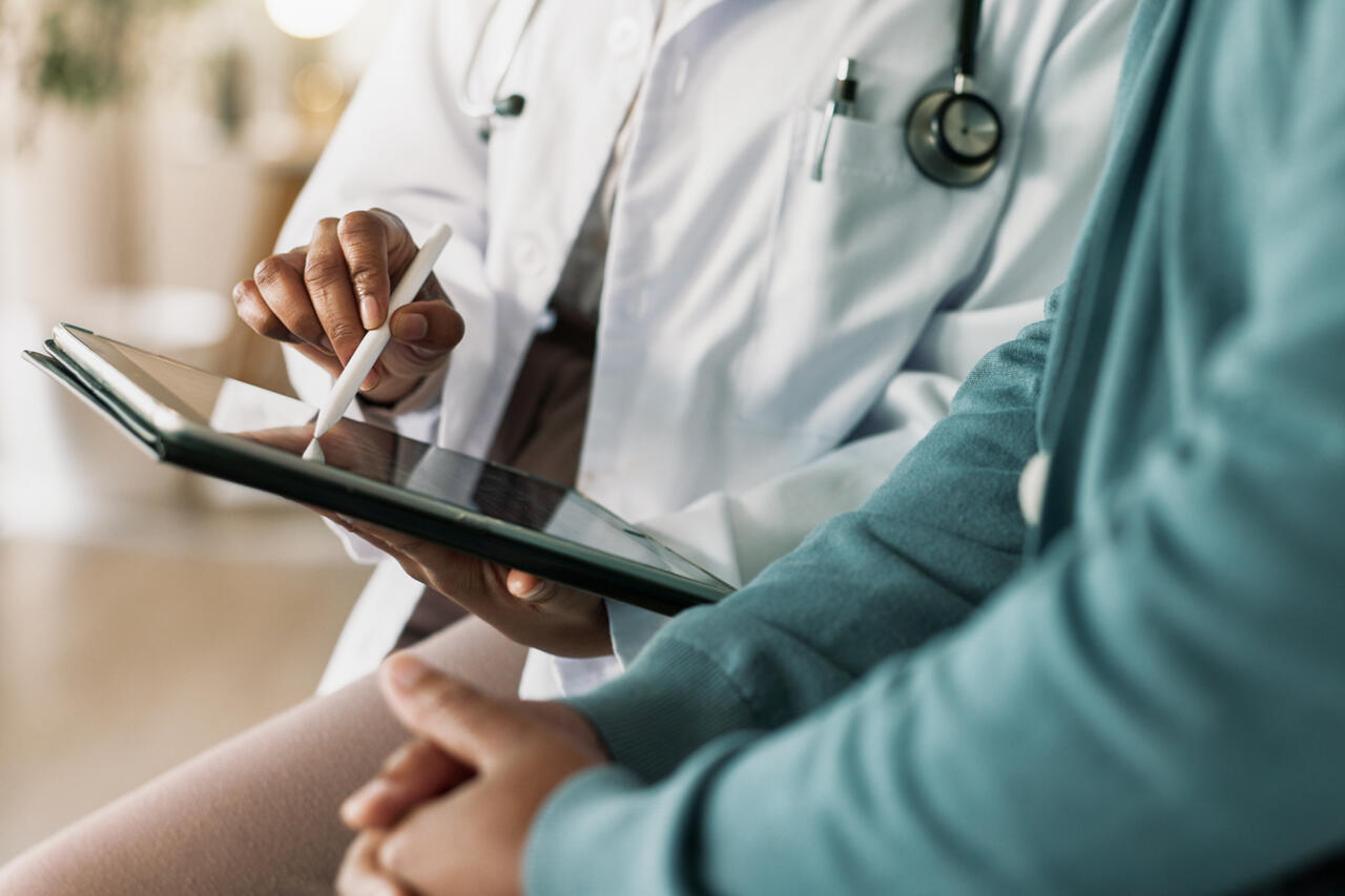 A close-up view of a unidentifiable doctor holding a stylus to tap on a tablet next to patient during a consultation.
