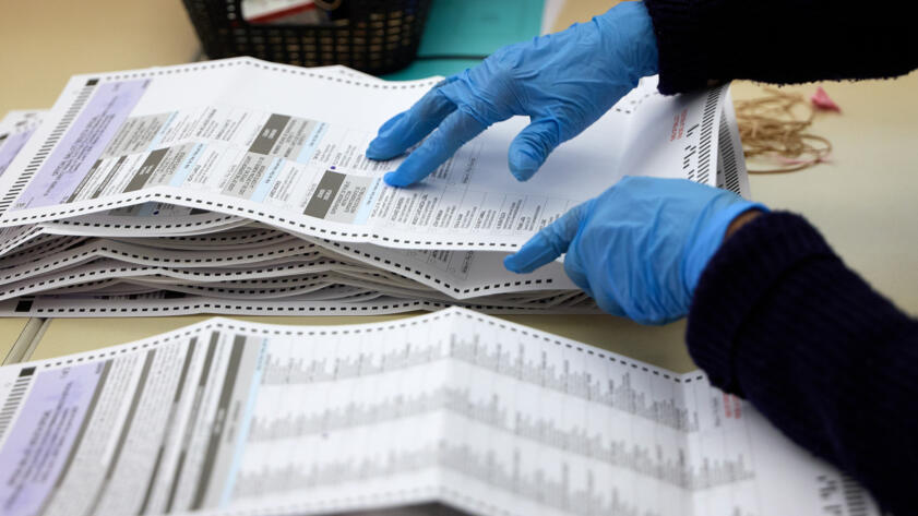 Blue-gloved hands sort and review stacks of election ballots on a table. The unfolded ballots display candidate races and ballot measures, while multiple sheets are organized into piles during the vote-counting process.